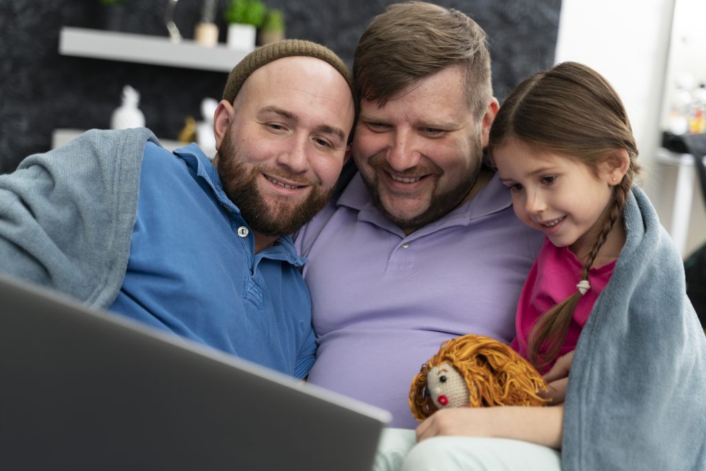 A happy family of two dads and their young daughter sitting together, smiling as they look at a laptop. This image represents the importance of family support during legal processes, such as navigating probate in Illinois, with compassionate legal guidance."